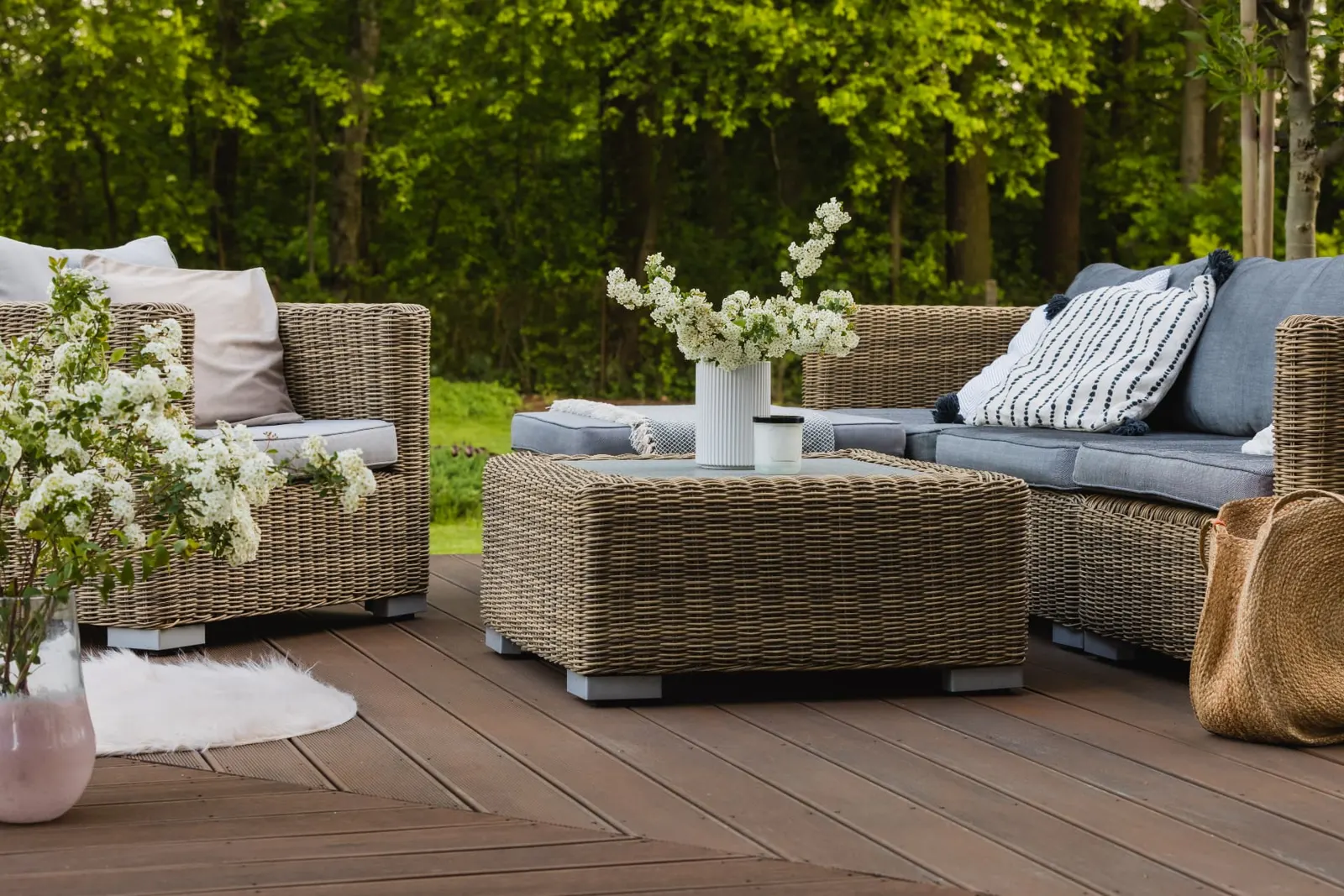 A garden deck with wicker lounge seating, soft grey cushions and white blossom against green trees
